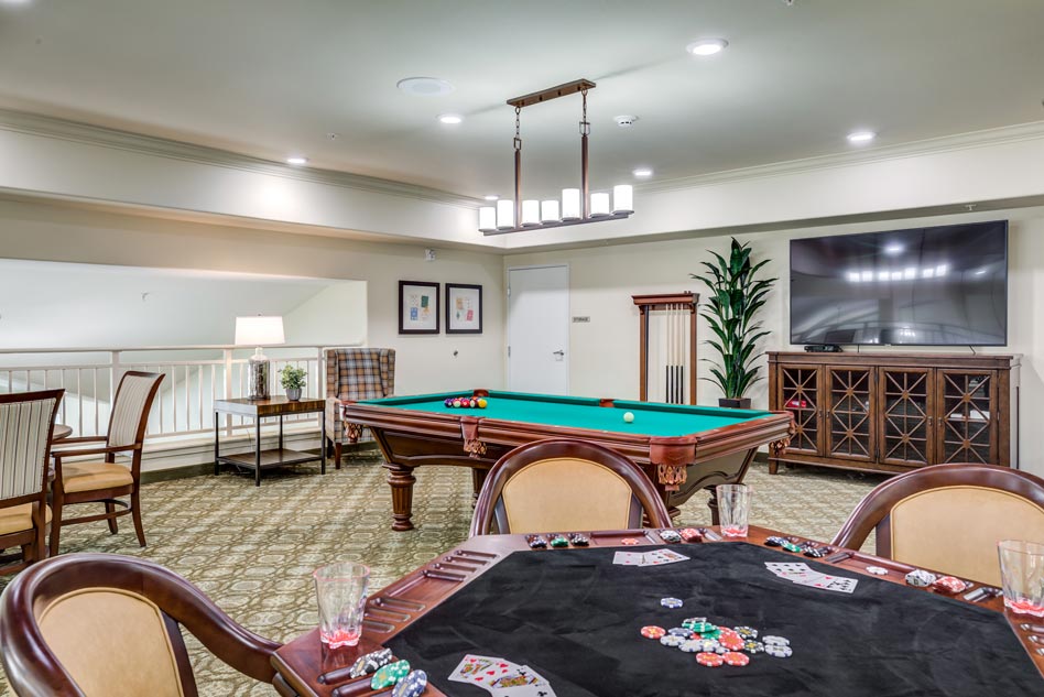 A game room with a green-felt pool table, poker table with cards and chips, wooden chairs, a flat-screen TV, two framed pictures on the wall, a potted plant, and a wooden cabinet. The room has beige walls and carpet flooring with a green and brown pattern.