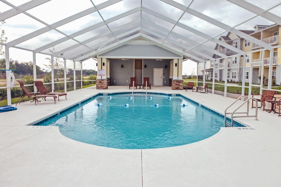 An indoor swimming pool under a transparent roof with lounge chairs and tables around it. The pool has a ladder on one side, floating lane dividers, and steps for entry. Buildings and grassy areas are visible in the background outside the enclosed structure.