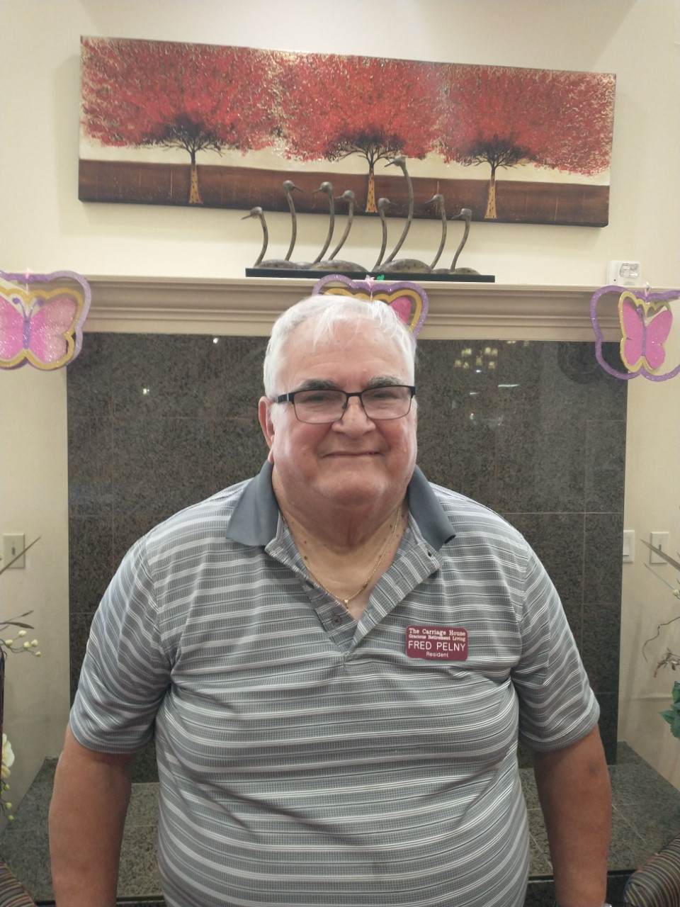 A man with white hair and glasses, wearing a gray striped polo shirt with a name tag, stands smiling in front of a dark marble wall. The wall is decorated with purple butterfly ornaments and has a colorful painting of red trees on it.