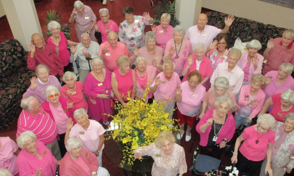 A large group of elderly people, mostly women, are gathered in a circular formation indoors. They are all wearing various shades of pink clothing and are smiling and waving at the camera. A table with a large arrangement of yellow flowers is in the center.