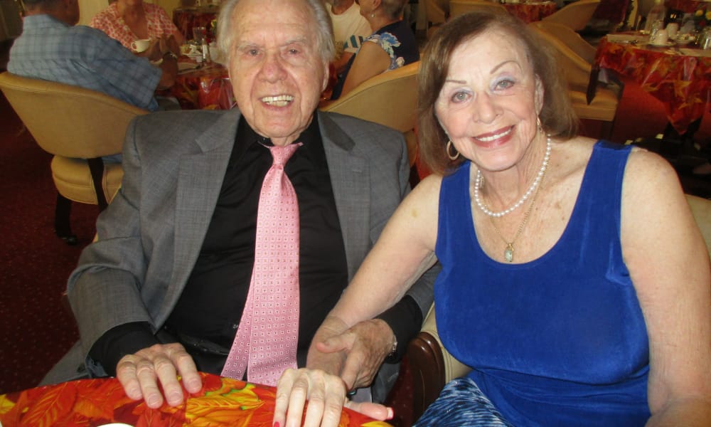 An elderly man in a suit and tie sits beside an elderly woman in a sleeveless blue dress and pearl necklace. They are smiling and sitting at a table with a bright, colorful tablecloth. Other people are visible in the background, seated at tables in the same setting.