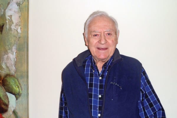An elderly man with short white hair smiles at the camera. He is wearing a blue and black plaid shirt with a dark blue vest over it. Behind him, to the left, is a portion of a painting with green, white, and red colors. The background wall is light-colored.