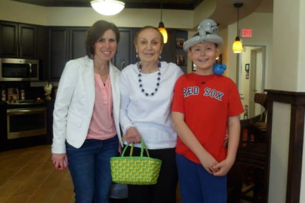 A woman in a white blazer, an elderly woman holding a green basket, and a boy in a red "Red Sox" shirt with a balloon hat stand together in a room with dark cabinets and wooden flooring. The boy has a smile on his face, and the room is well-lit.