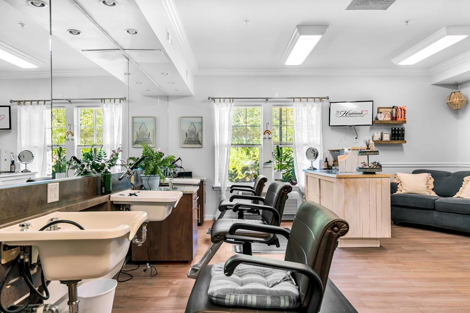 This image shows a well-lit, modern salon with large windows and plenty of natural light. There are two black salon chairs, mirrors, sinks, and plants. A seating area with a couch and decor items are visible in the background. The overall atmosphere is inviting and cozy.