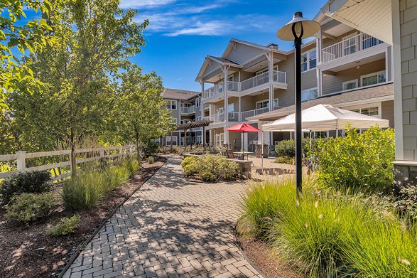 A pathway lined with greenery leads to a multi-story residential building. The building features balconies and large windows. Outdoor patio tables are shaded by red and white umbrellas. A lamppost stands on the right side, with a bright blue sky above.