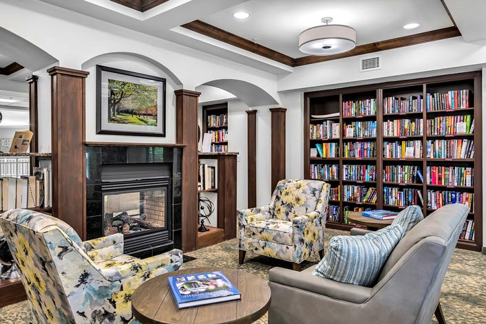 A cozy library room with bookshelves filled with colorful books, a fireplace, and three patterned armchairs arranged around a round wooden table. Bright lighting illuminates the space, and a framed picture hangs above the fireplace.