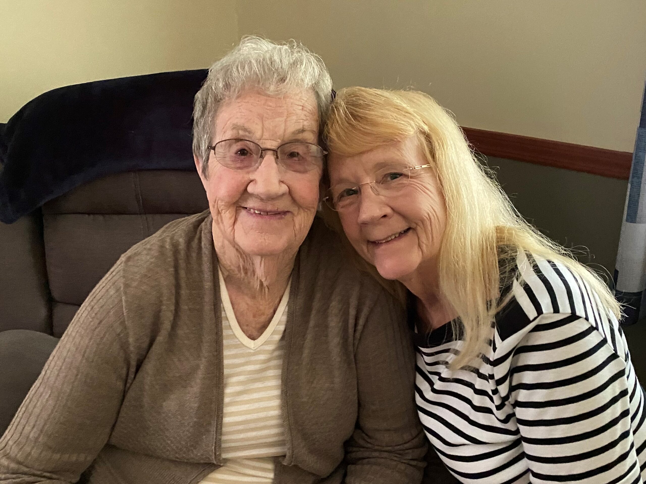 Two elderly women are seated, smiling warmly at the camera. One has short, curly gray hair and wears glasses, a light striped shirt, and a beige cardigan. The other has long blond hair and also wears glasses, with a black and white striped shirt. They are sitting closely together.