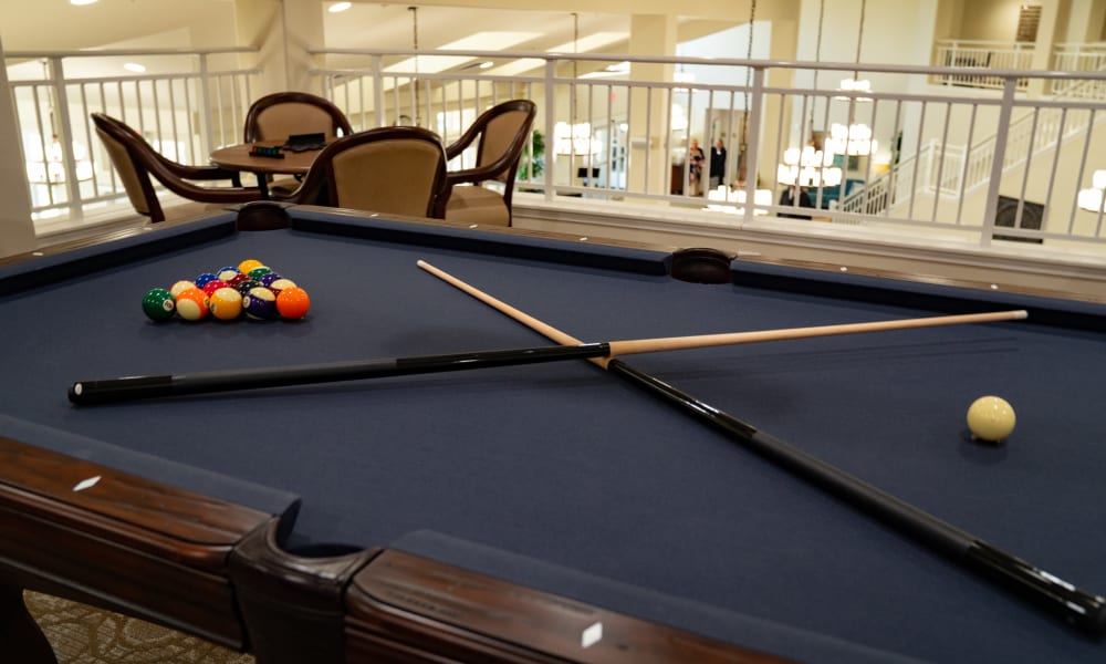 A pool table with a complete set of racked balls, two cues placed in an X shape, and a cue ball ready for a break shot. In the background, there are chairs and a small table under soft lighting, and a staircase visible beyond the railing.