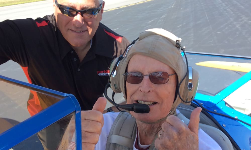 An elderly man wearing aviator headphones and a pilot cap gives a thumbs-up while sitting in the cockpit of a small airplane. A man wearing sunglasses and a black shirt with red stripes stands beside the open cockpit door, also giving a thumbs-up.
