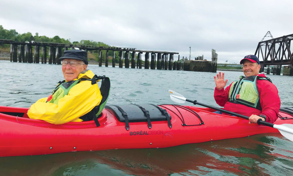 Two people are kayaking in red kayaks on a cloudy day near an old bridge structure. The person in the front kayak, wearing a yellow jacket and black cap, smiles at the camera. The person in the rear kayak, wearing a red jacket and black cap, waves cheerfully.