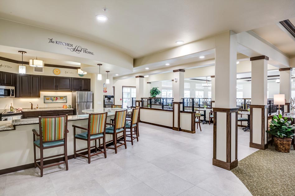 A spacious, well-lit common area features a modern kitchen with wooden cabinets, stainless steel appliances, and a long counter with colorful cushioned bar stools. The adjacent dining area has several tables and chairs, with plants and large windows adding to the ambiance.