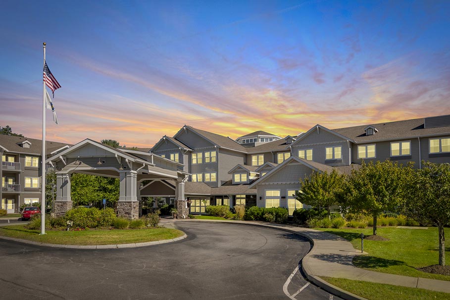 A large, three-story building with gray siding and white trim stands under a colorful sunset sky. The building features multiple windows and a covered entrance with a roundabout driveway. A flagpole displaying flags is located on the left side of the driveway.