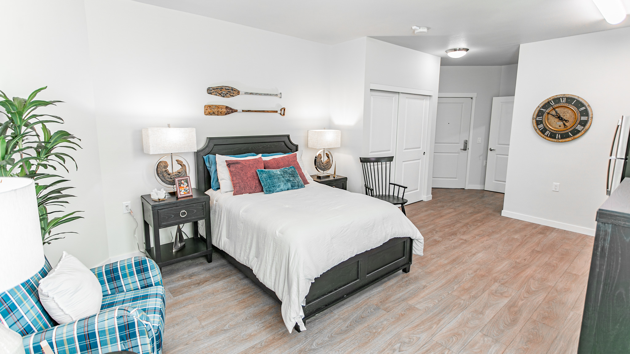 A cozy, modern bedroom with a black bed frame, white bedding, colorful pillows, two lamps on nightstands, a plaid armchair, a large wall clock, and wooden paddles as wall decor. Light wood floors and white walls.