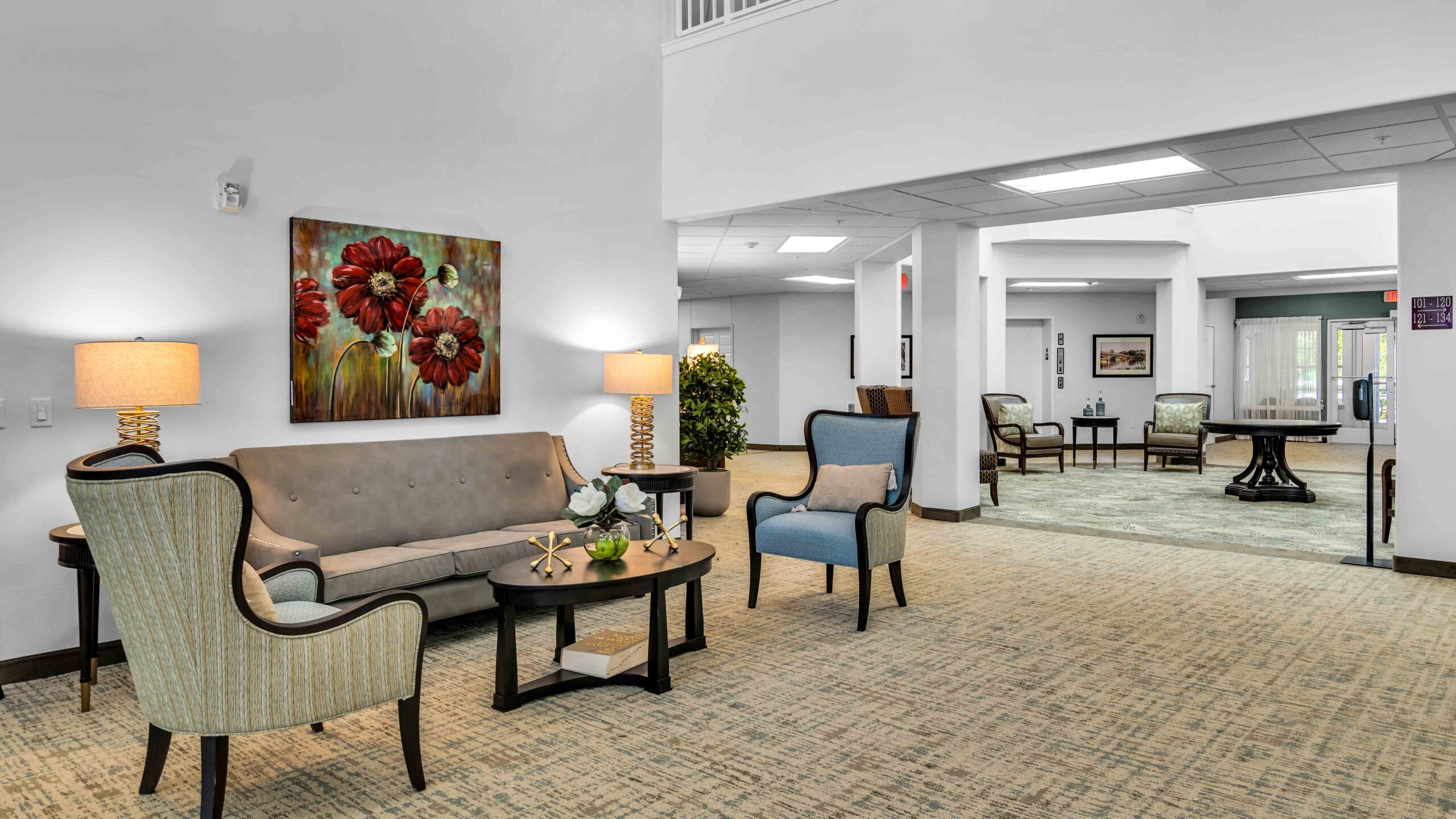 A bright, spacious lobby with a beige sofa, two armchairs, coffee tables, table lamps, and a large floral painting on the wall. Additional seating is visible in an adjoining room through a wide doorway.