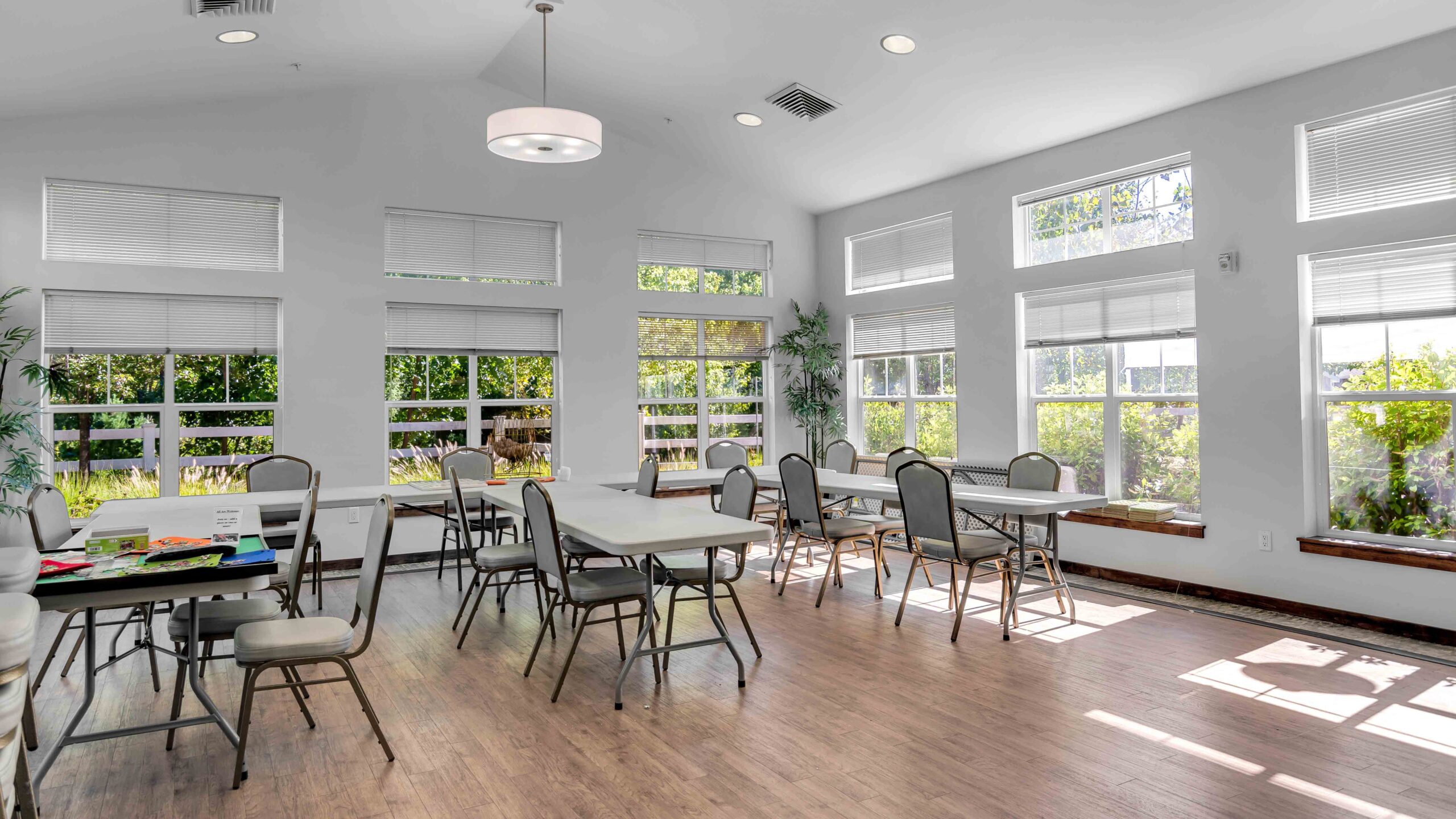 A bright room with large windows, several tables and chairs, and sunlight streaming in. The space is arranged for group activities, with games and puzzles visible on one table. Greenery is visible outside the windows.