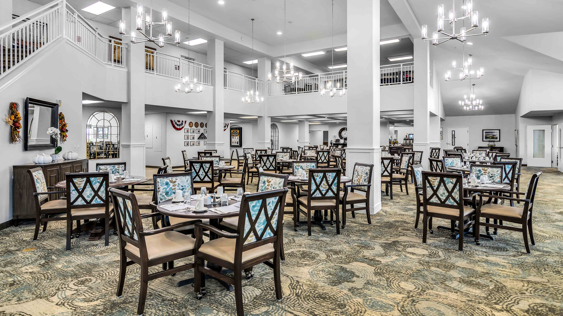 Spacious, elegant dining room with multiple tables and chairs, place settings, chandeliers, large windows, patterned carpet, and high ceilings, creating a bright and welcoming atmosphere.