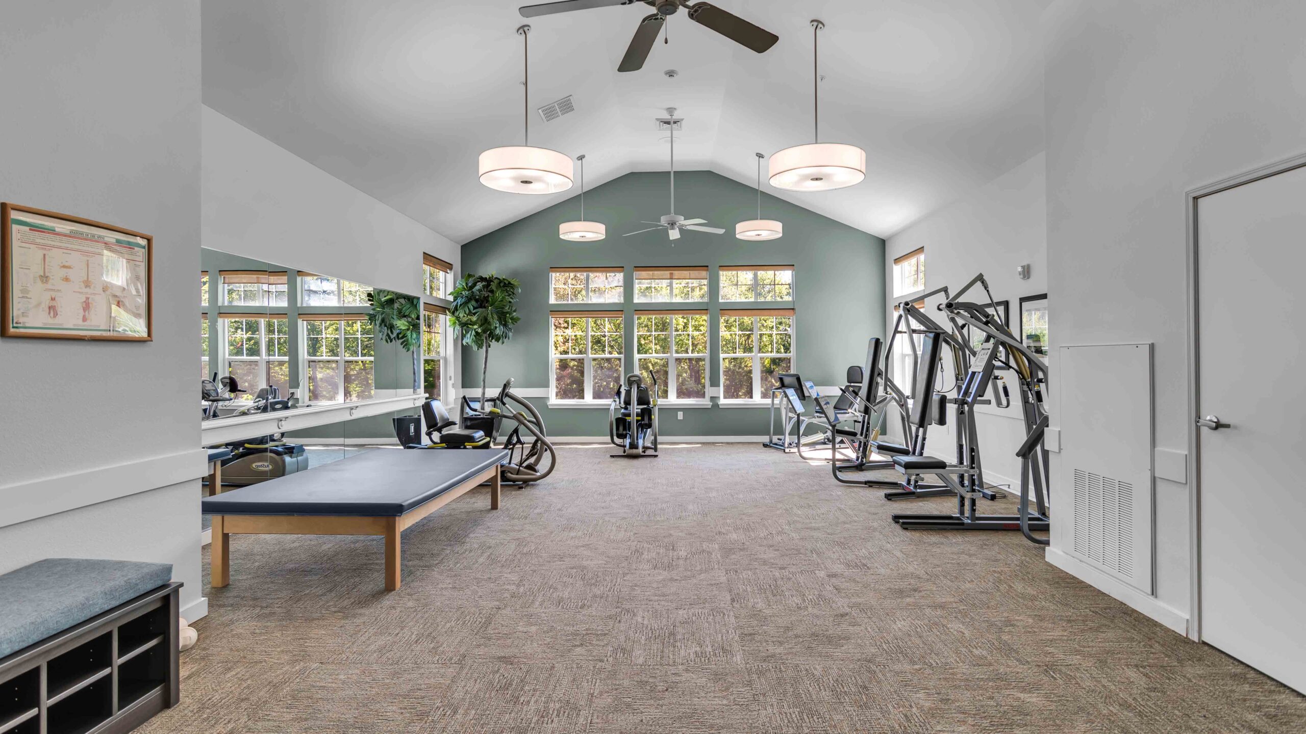 A spacious, well-lit gym with large windows, exercise machines, a padded table, mirrors along one wall, and ceiling fans. The room has neutral colors and ample space for workouts.