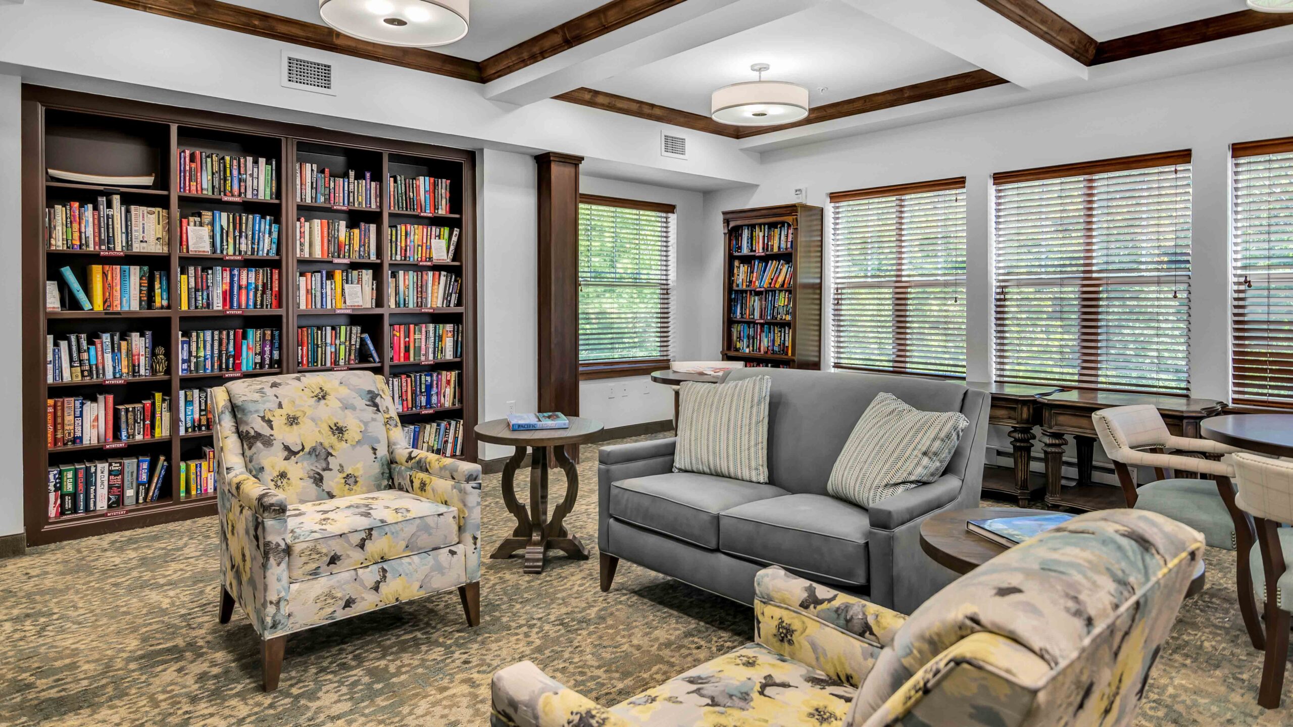 Cozy library room with bookshelves, two floral armchairs, a gray sofa, side tables, and large windows with blinds letting in natural light. The room features carpeted flooring and wooden ceiling beams.