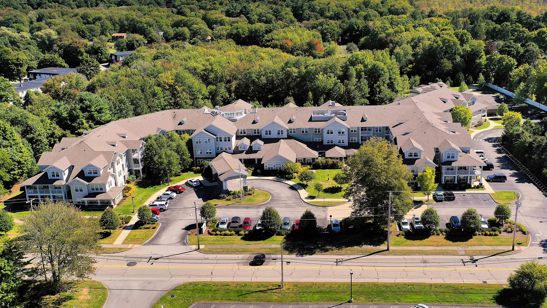 Aerial view of a large, light-colored residential building complex with multiple wings, surrounded by trees and greenery, with cars parked in front and a road running along the front of the property.