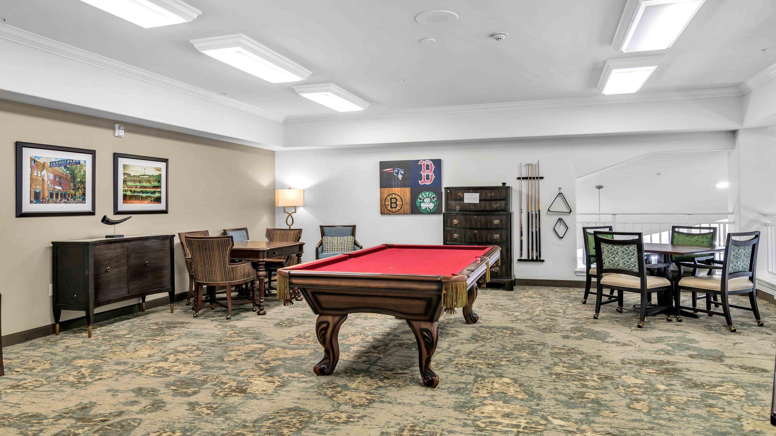 A game room with a red pool table, two round tables with chairs, wall art, a lamp, and a cue rack. The space is well-lit with fluorescent ceiling lights and has patterned carpet flooring.