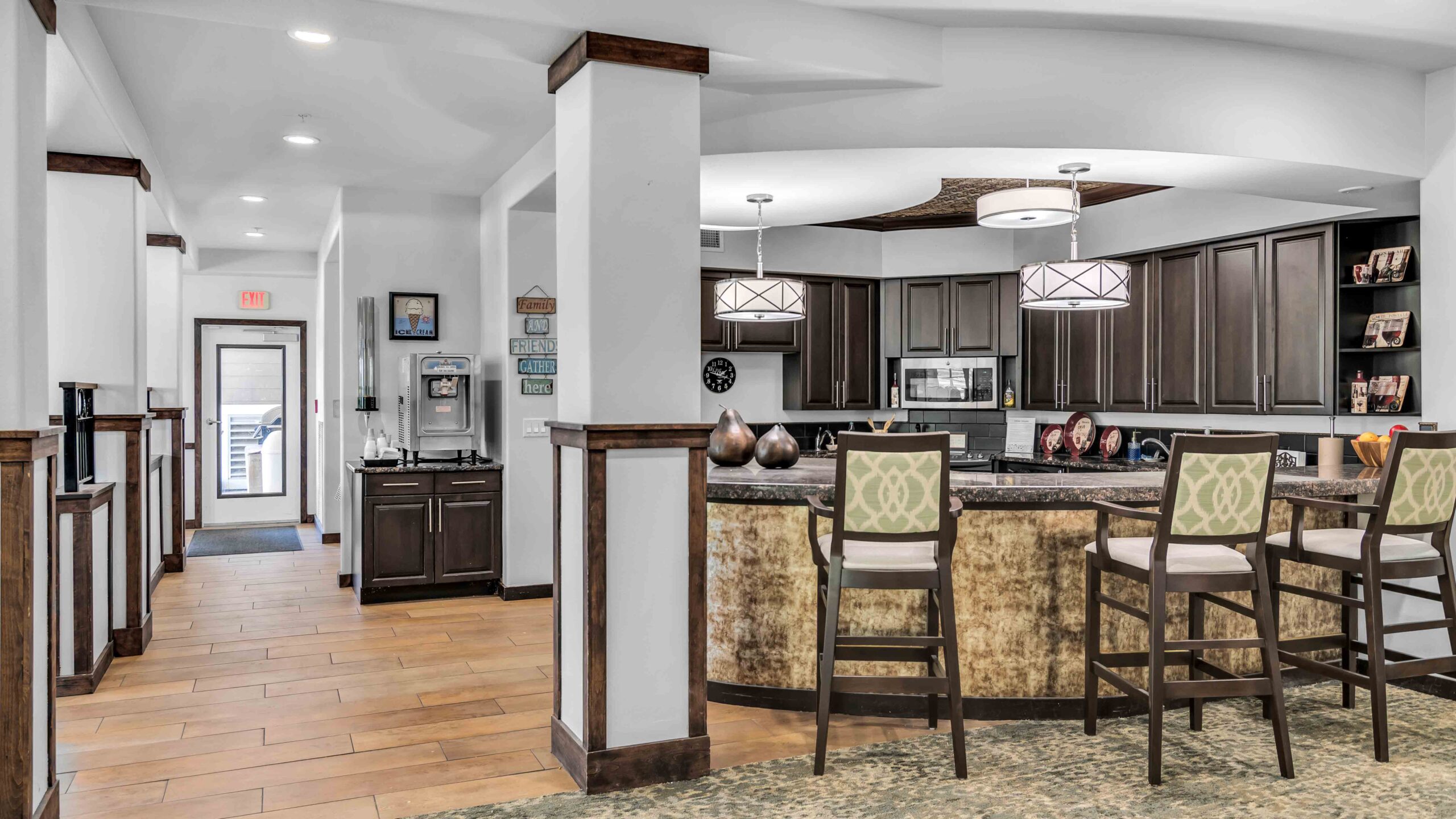 Modern kitchen with dark cabinets, a large island with barstools, patterned carpet, and wood accents; hallway to the left with artwork and wall sconces, and bright lighting throughout the space.