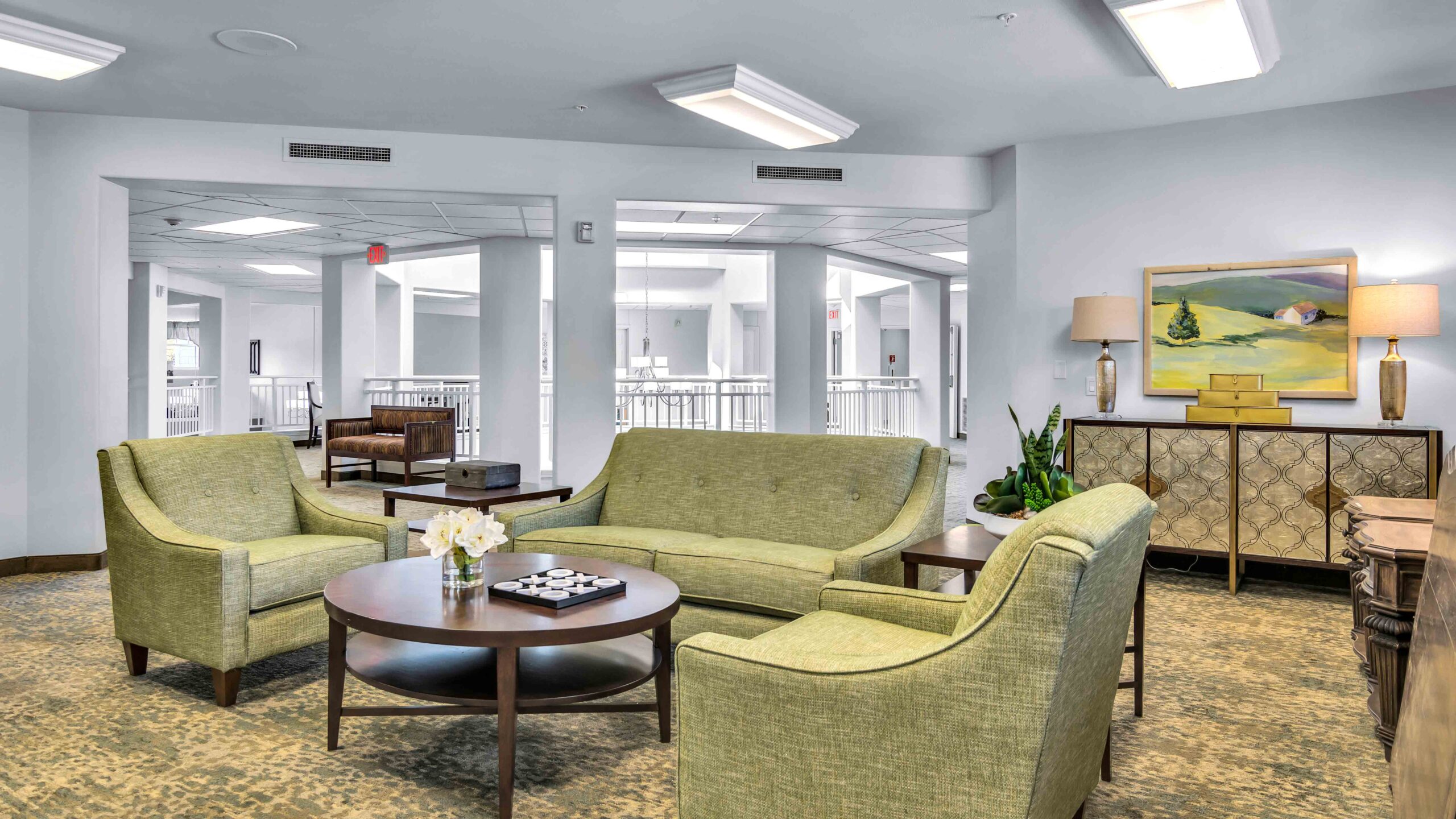 A modern lounge area with green upholstered sofas and armchairs around a round coffee table, set on a patterned carpet. In the background, a sideboard, lamps, and a landscape painting adorn the wall.