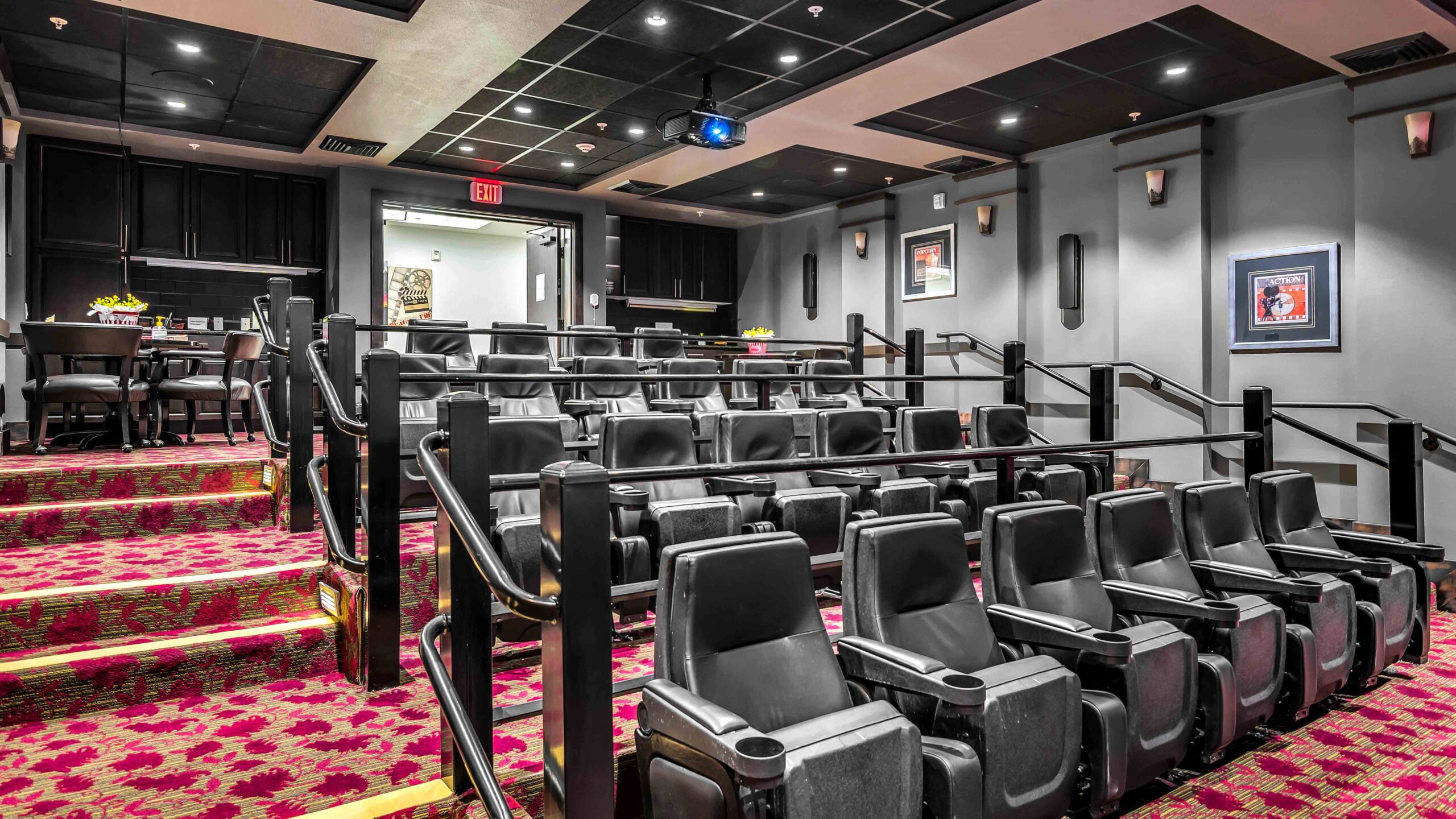 A small, modern theater room with tiered rows of black cushioned seats, red patterned carpet, and a projector on the ceiling. The walls are gray with framed art, and a lounge area is visible in the back.