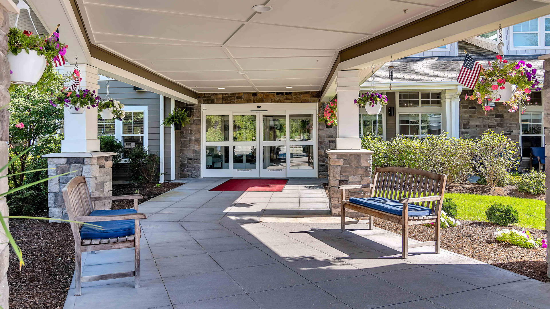 Covered entrance to a building with hanging flower baskets, benches on both sides, a red welcome mat, glass doors, and landscaped greenery surrounding the walkway.