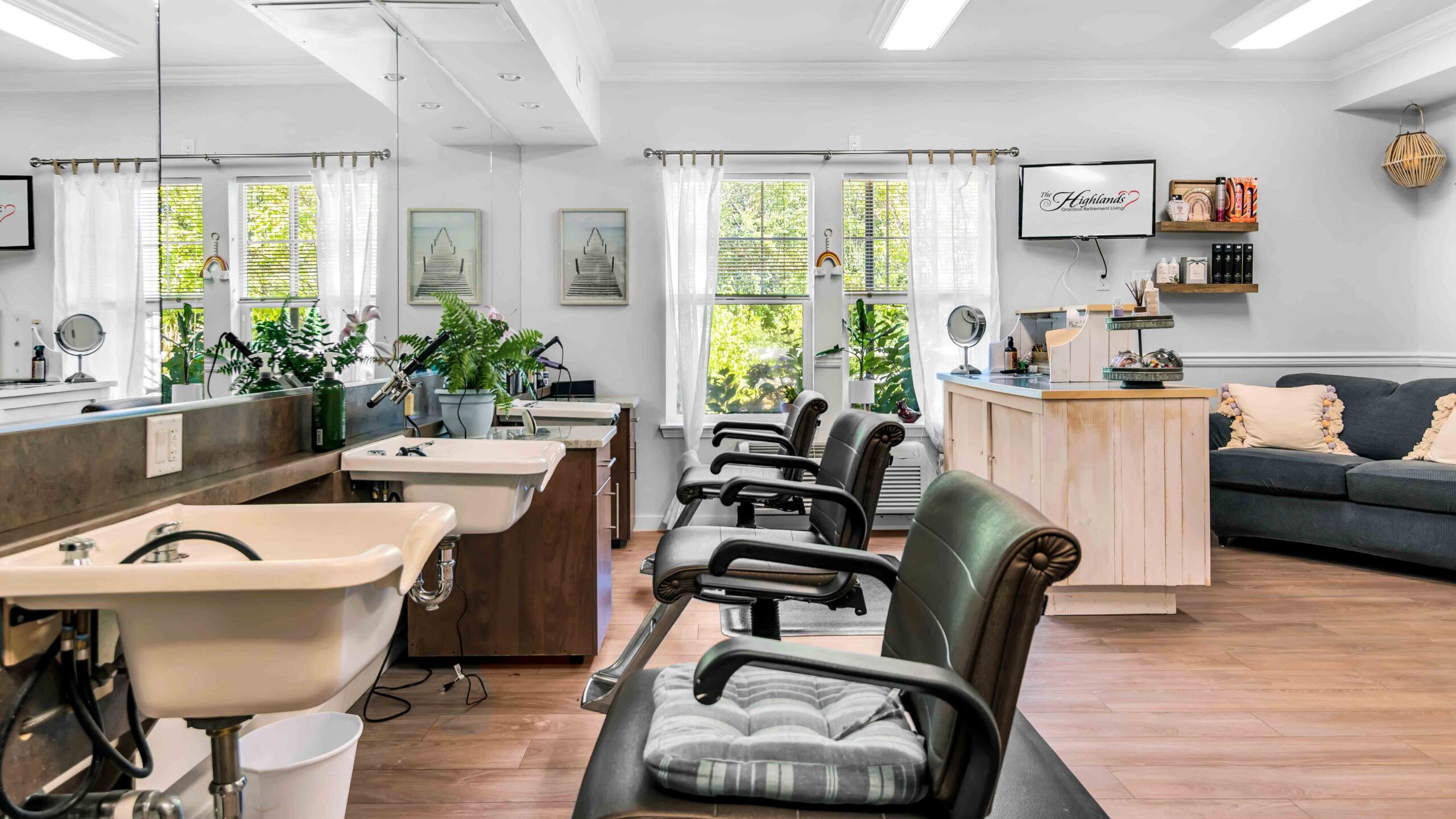 A bright, inviting hair salon with three black chairs, washbasins, a wooden reception desk, houseplants, wall art, shelves with products, and large windows letting in natural light.