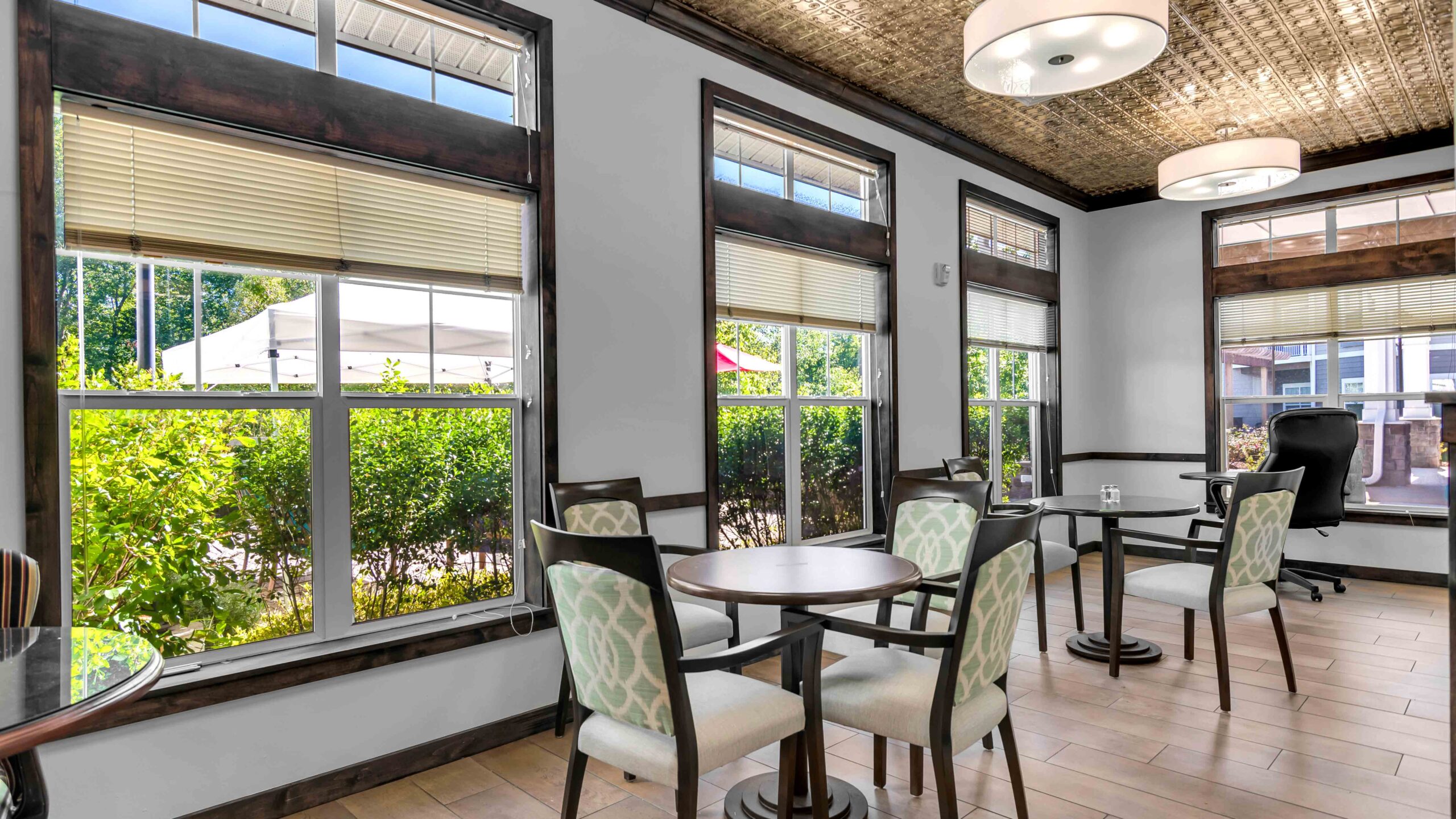 A bright room with large windows, wooden floors, and several small round tables with cushioned chairs. Sunlight streams in, and greenery is visible outside the windows. The ceiling features decorative lighting.