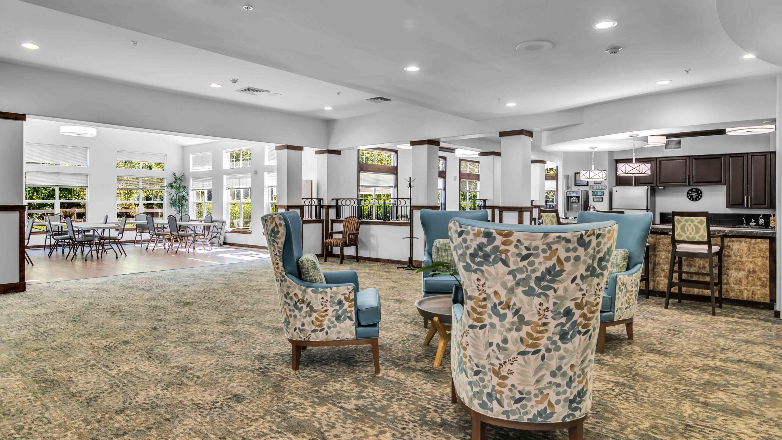 Spacious, well-lit lounge area with patterned armchairs, carpeted floor, and large windows. Dining tables and chairs are visible in the background, along with a kitchenette on the right.
