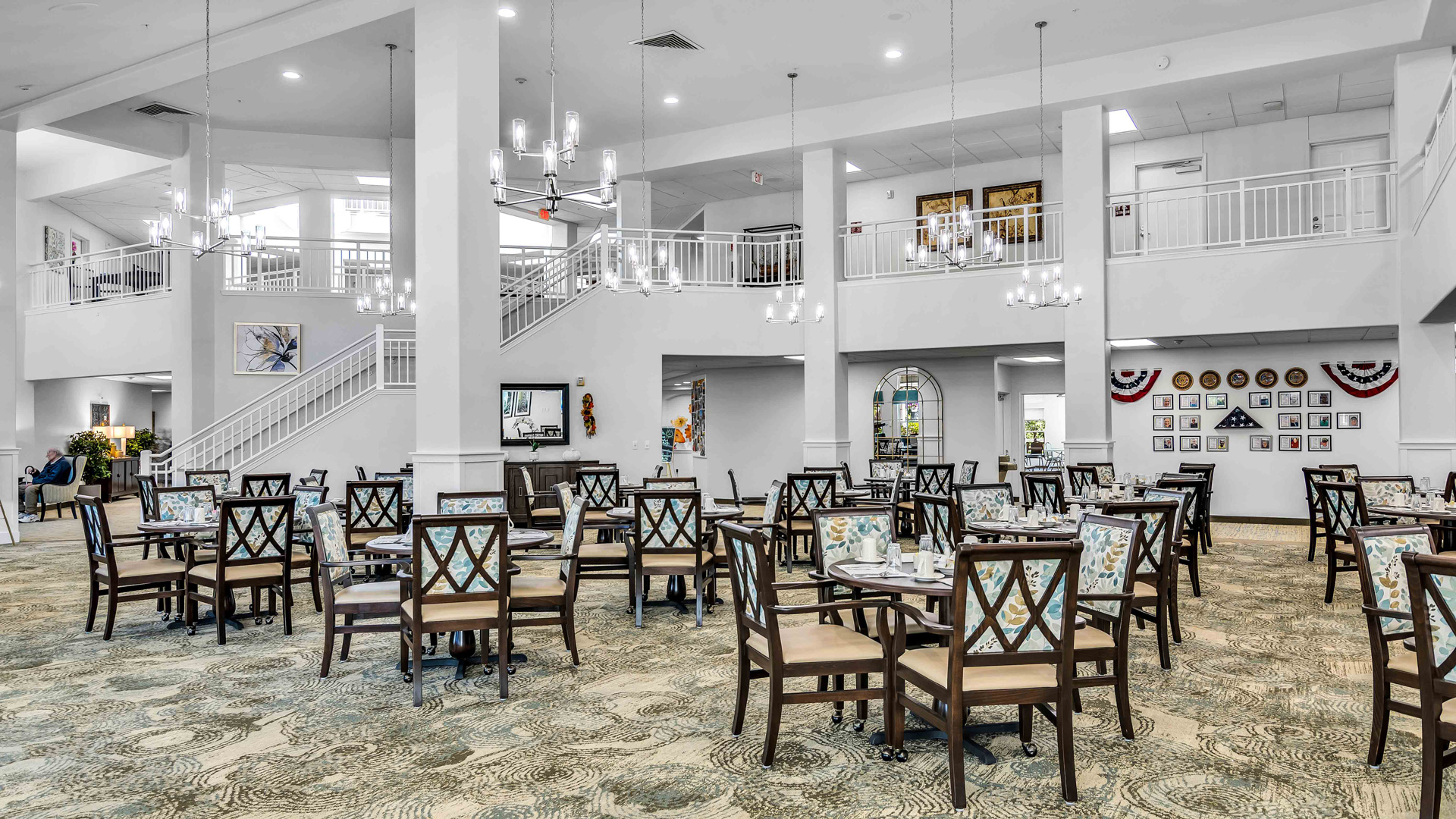 A spacious, elegant dining room with multiple tables and chairs, high ceilings, chandeliers, and large windows. White walls, decorative art, and a staircase leading to an upper balcony are visible.