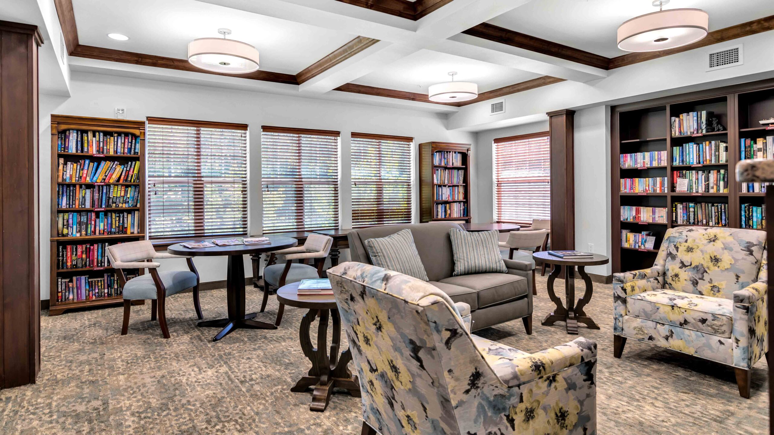 A cozy library room with bookshelves filled with books, several round tables with chairs, armchairs with floral patterns, a sofa, and large windows letting in natural light.