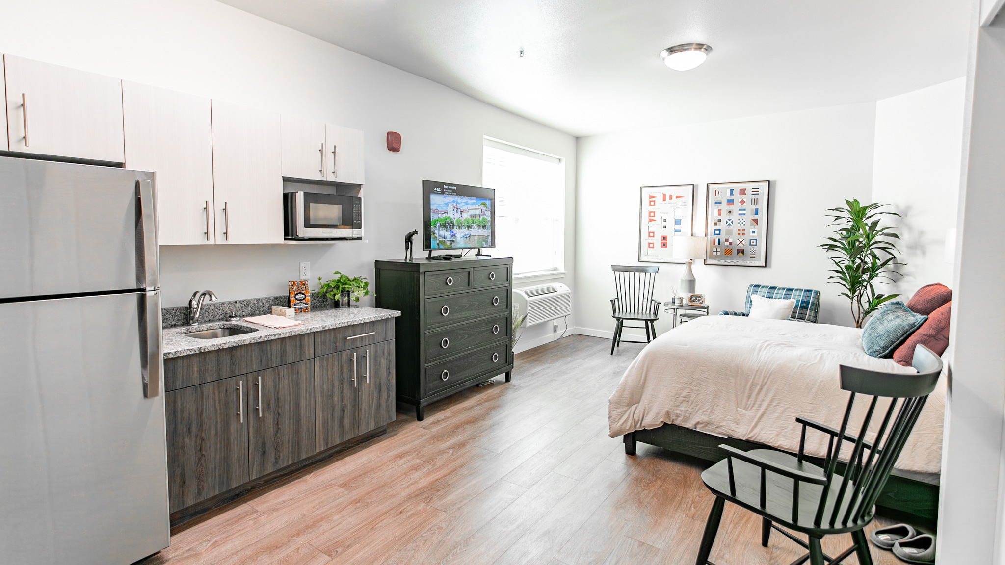 A modern studio apartment features a kitchenette with stainless steel appliances, a TV atop a black dresser, a bed with colorful pillows, wooden chairs, wall art, and a large window letting in natural light.