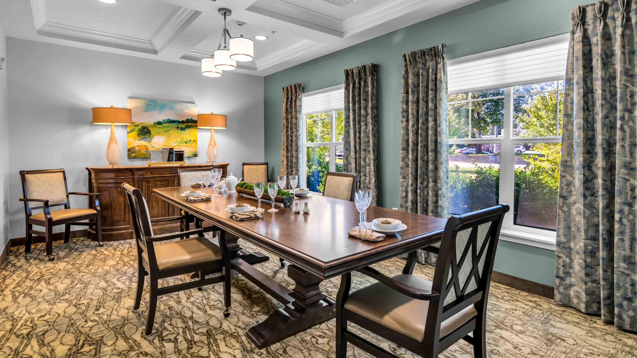 A bright, elegant dining room with a large wooden table set for six, upholstered chairs, a sideboard with lamps and artwork, patterned curtains, and large windows letting in natural light.