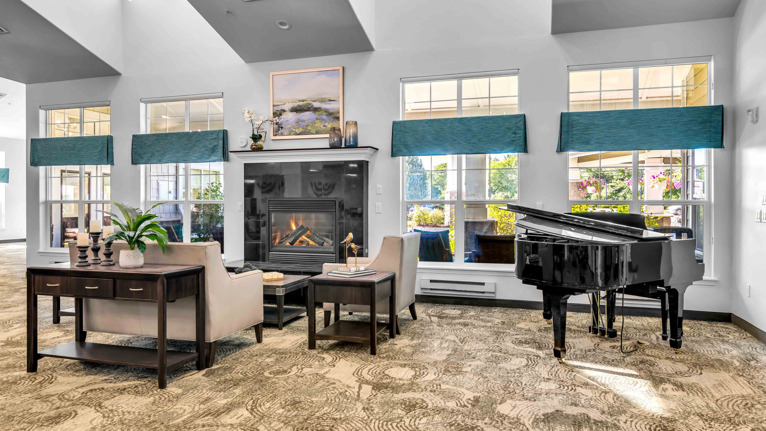 A bright, elegant living room with large windows, a fireplace, two beige armchairs, a black grand piano, and decorative accents, including plants, candles, and artwork above the mantel.