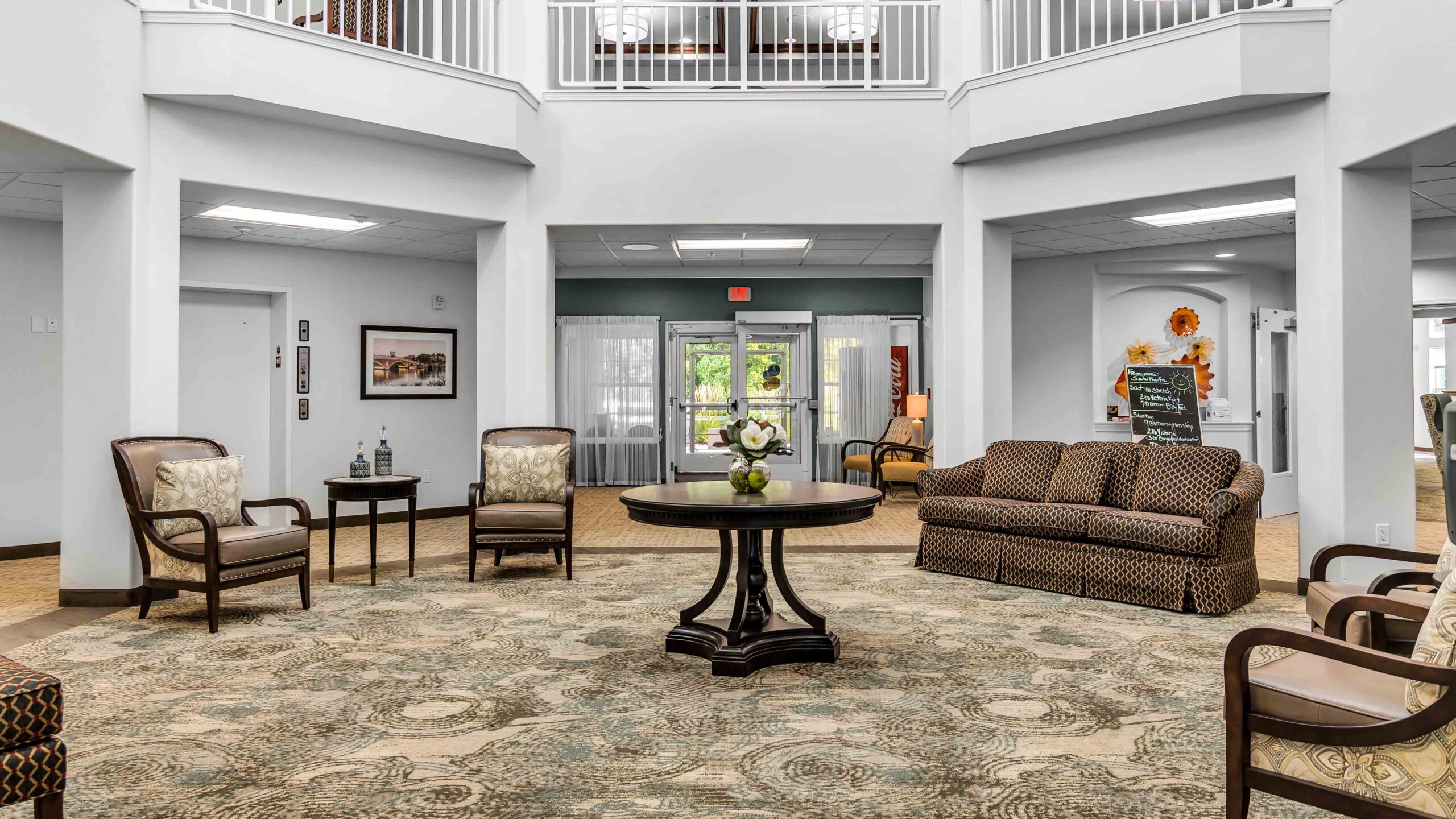 A spacious, well-lit lobby with a patterned rug, round table with a floral centerpiece, cushioned chairs, and a sofa. Large windows and glass doors let in natural light; framed art decorates the walls.