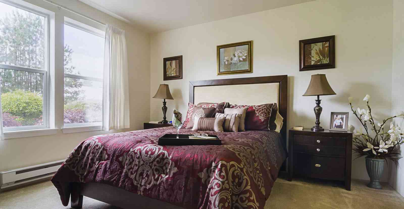 A cozy bedroom with a queen-sized bed covered in a red and beige patterned comforter set. There are nightstands with lamps on both sides of the bed, and framed artwork on the walls. A large window with white curtains allows natural light to fill the room.