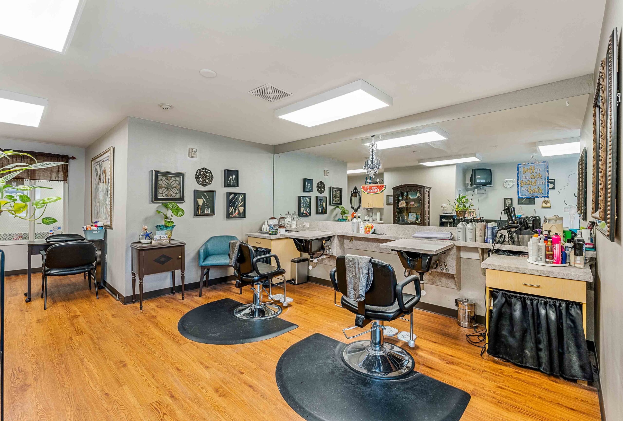 A bright and tidy hair salon with light gray walls, wooden flooring, and large mirrors. Two black salon chairs face individual workstations equipped with hair styling tools and products. Wall decorations and plants add a cozy touch. Multiple light fixtures illuminate the room.