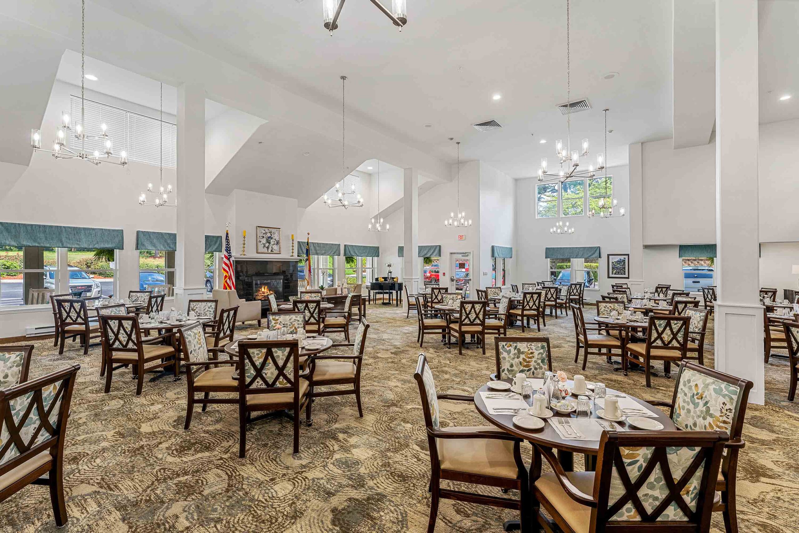 A spacious dining hall with multiple round tables set for meals. Each table has plates, cups, and floral centerpieces. Elegant chandeliers hang from a high ceiling. Large windows allow natural light to enter, and green curtains frame the windows.