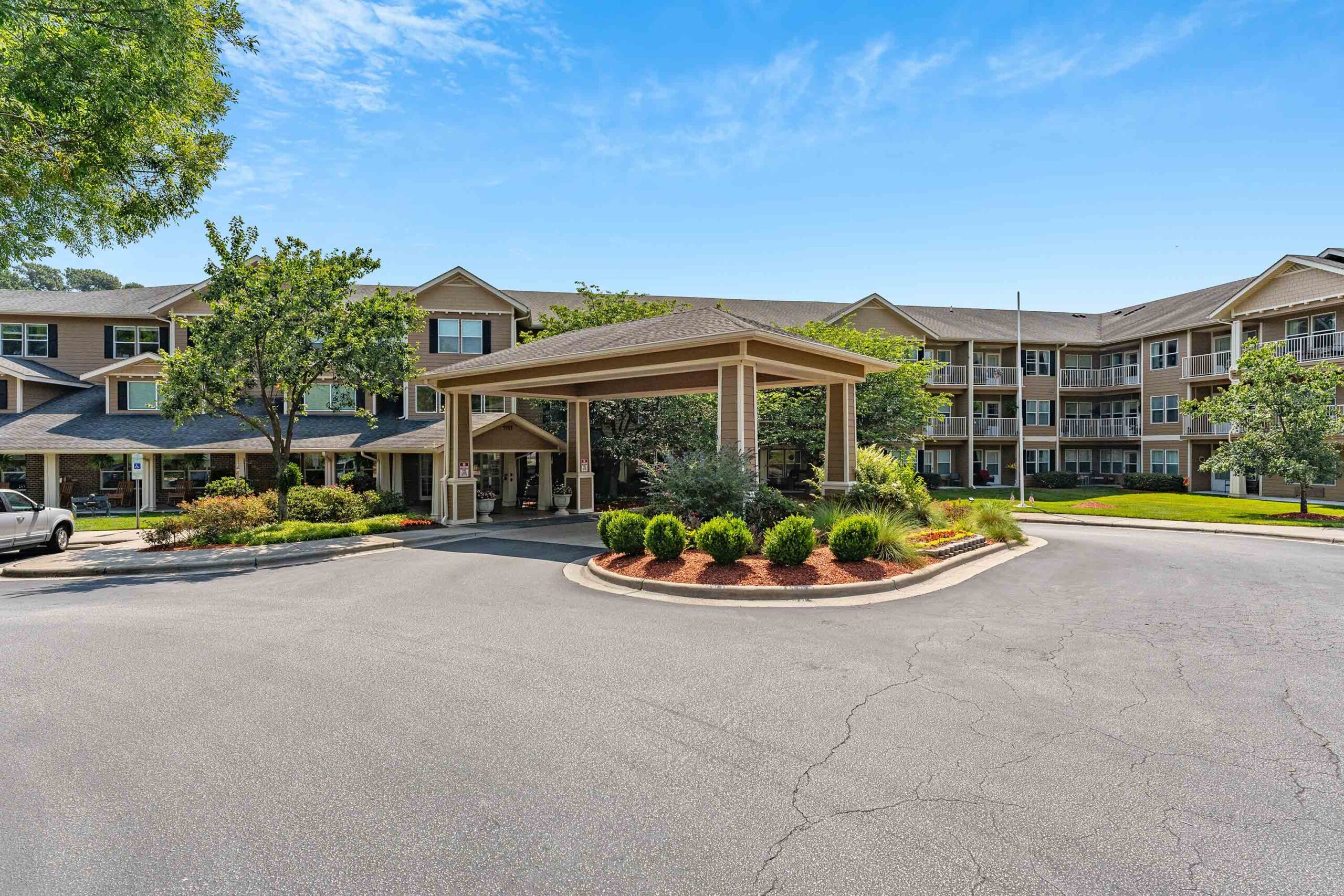 A large residential building with multiple floors is seen under a bright blue sky. A landscaped roundabout with shrubs and mulch is in front of the main entrance, which is covered by an extended canopy. A few trees and neatly trimmed bushes are alongside the building.