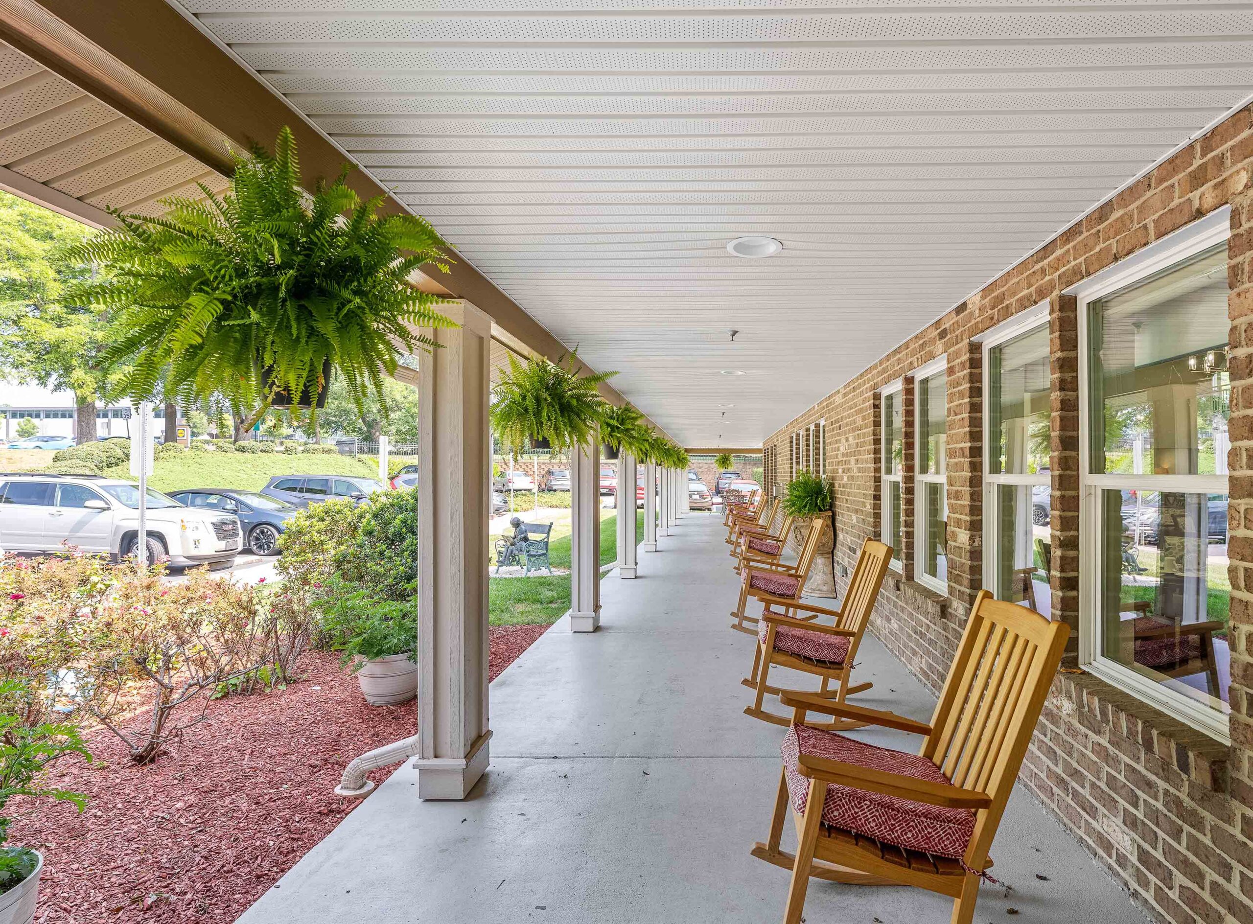 A long porch with wooden rocking chairs on one side and hanging ferns from the ceiling on the other. The porch is bordered by a red brick wall with several windows. Outside, there are parked cars and a well-maintained garden.