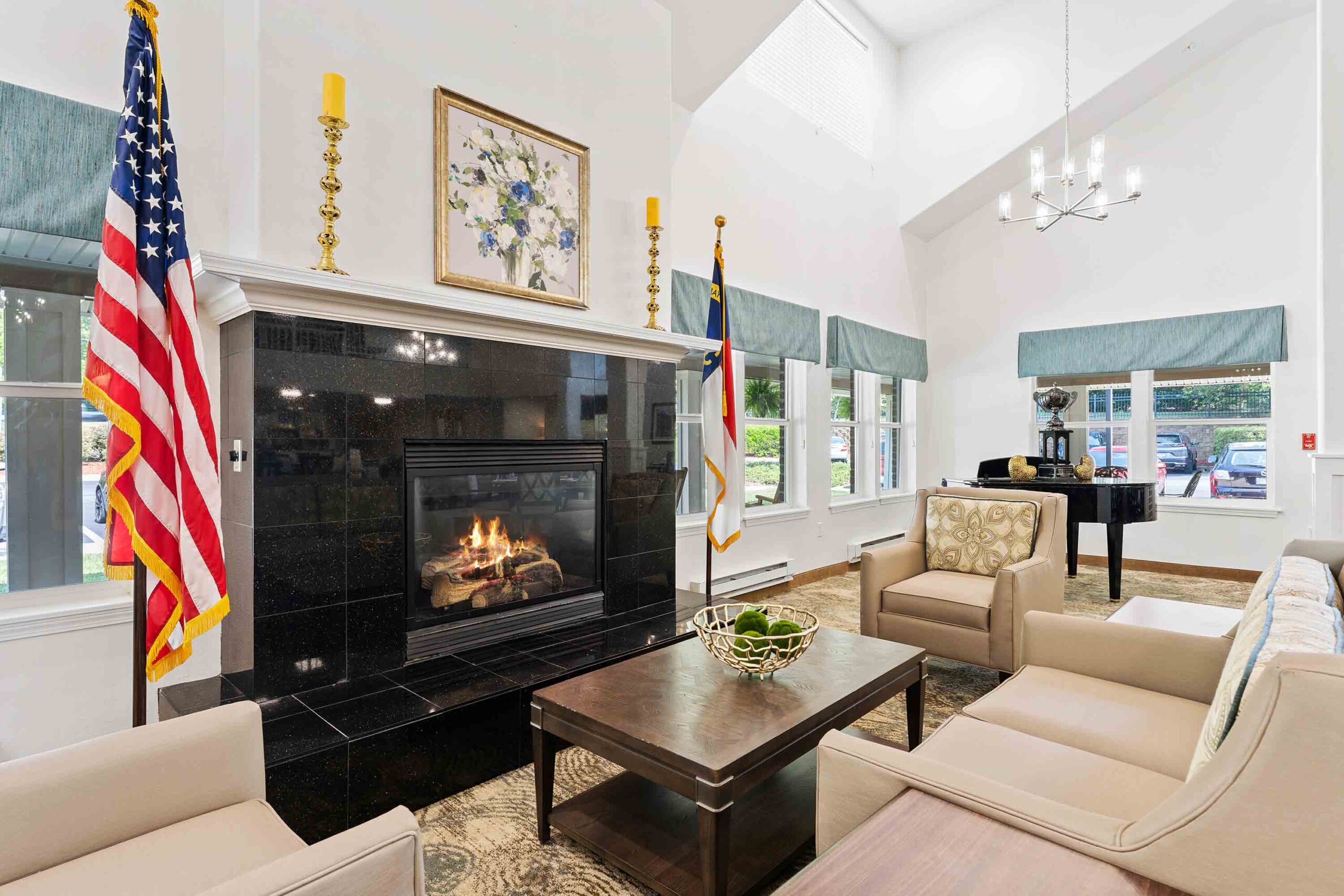 A cozy living room with a black marble fireplace, beige armchairs, and a wooden coffee table. Above the fireplace hangs a floral painting. American and state flags flank the fireplace. Windows with blue valances let in natural light, and a chandelier hangs from the high ceiling.