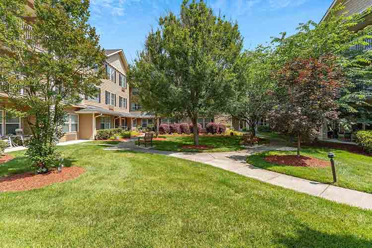 A well-maintained courtyard with green grass, shaded by various tall trees. Walkways lead to residential buildings on both sides. Benches and landscaped garden areas featuring shrubs and colorful flowers are scattered throughout the space. The sky is clear and blue.