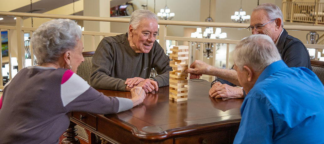 Four elderly people are gathered around a wooden table playing a game of Jenga. Three of them watch intently as one person carefully pulls a piece from the tower. They appear to be in a well-lit, comfortable indoor setting with chandeliers in the background.