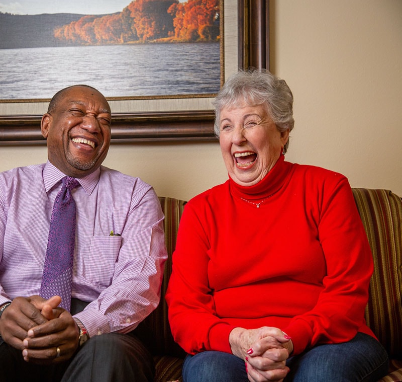 A man in a purple dress shirt and tie and a woman in a red sweater sit on a striped couch, laughing joyfully. A framed picture of a lake with autumn trees decorates the wall behind them.