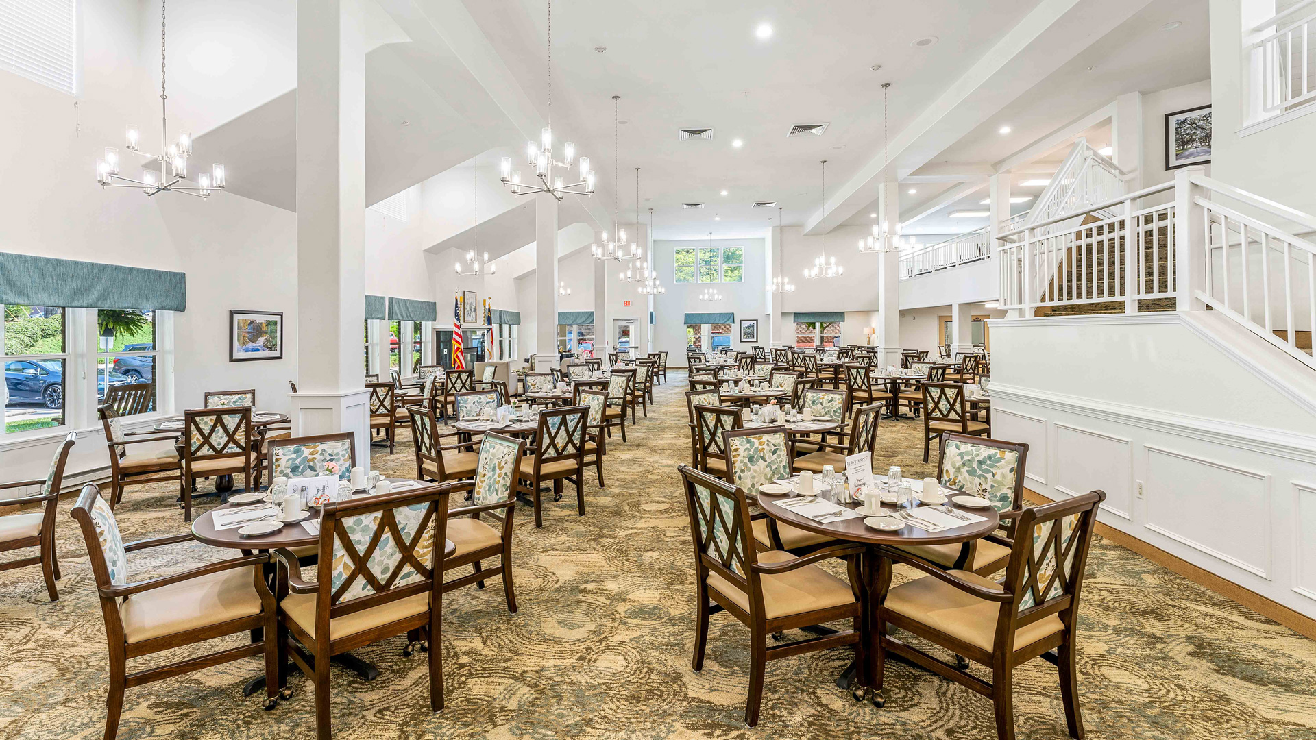 Spacious, elegant dining room with multiple round tables and chairs, set for a meal. Chandelier lights hang from the ceiling, and large windows provide natural light. A staircase is visible on the right.