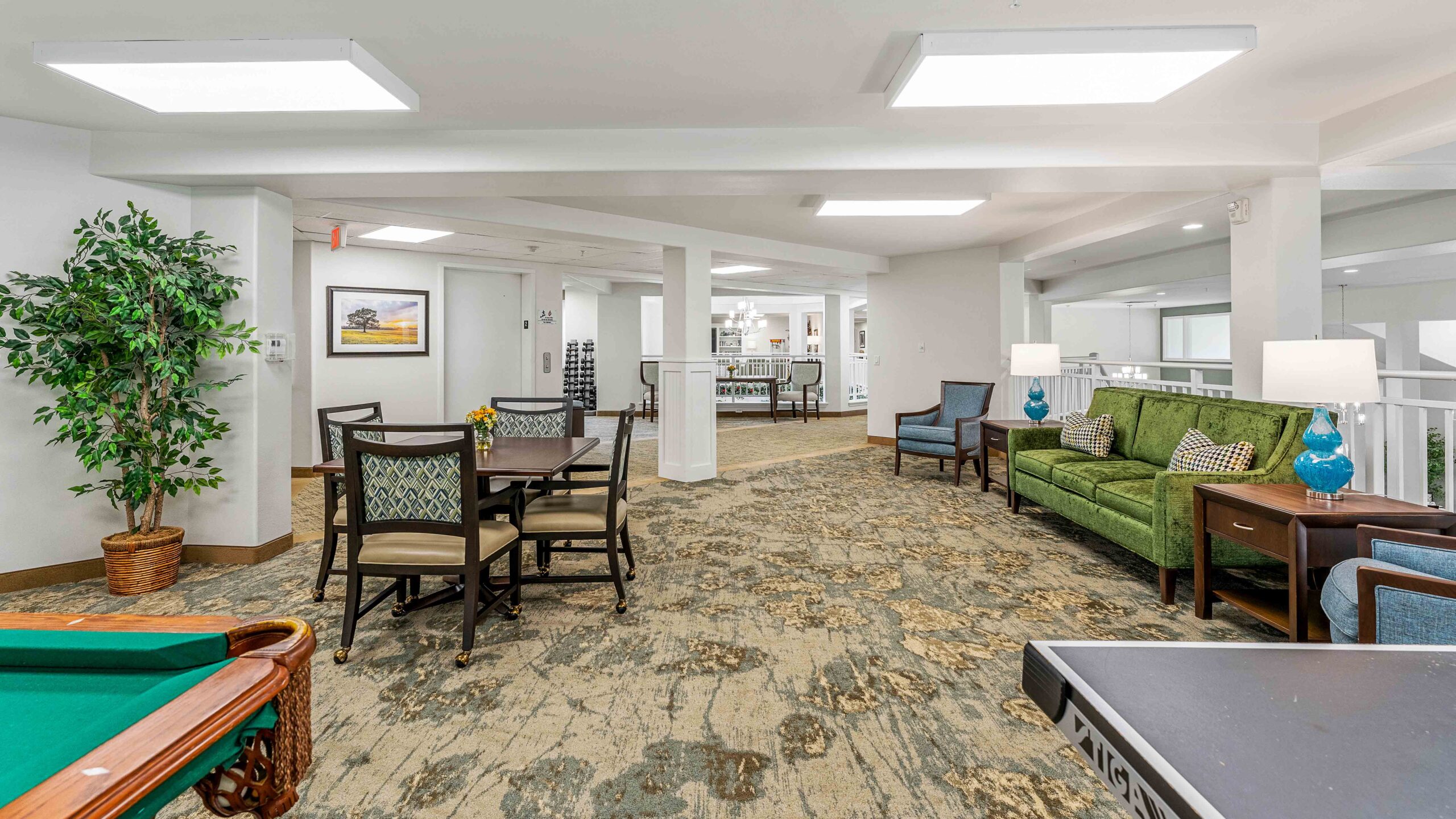 A spacious, well-lit lounge area features a green sofa, armchairs, a table with chairs, a pool table, potted plant, and decorative lamps, with neutral walls and carpeting, creating a cozy, inviting atmosphere.