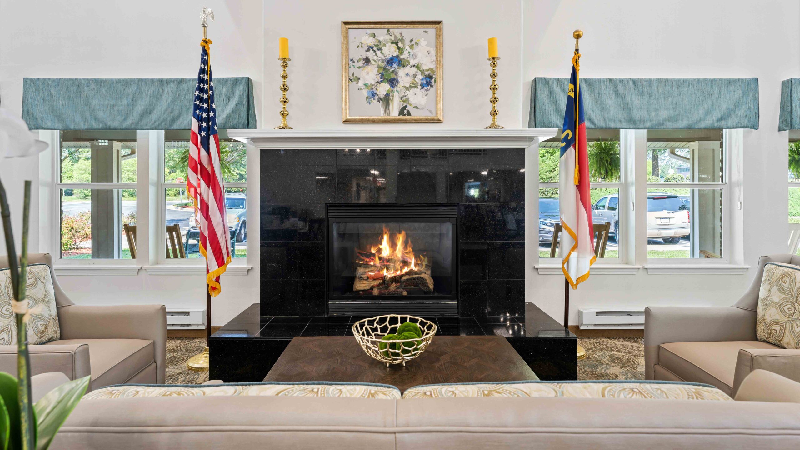 A cozy living room with a lit fireplace, two armchairs, a coffee table with a decorative bowl, and American and North Carolina flags on either side of the fireplace. Windows let in natural light on both sides.
