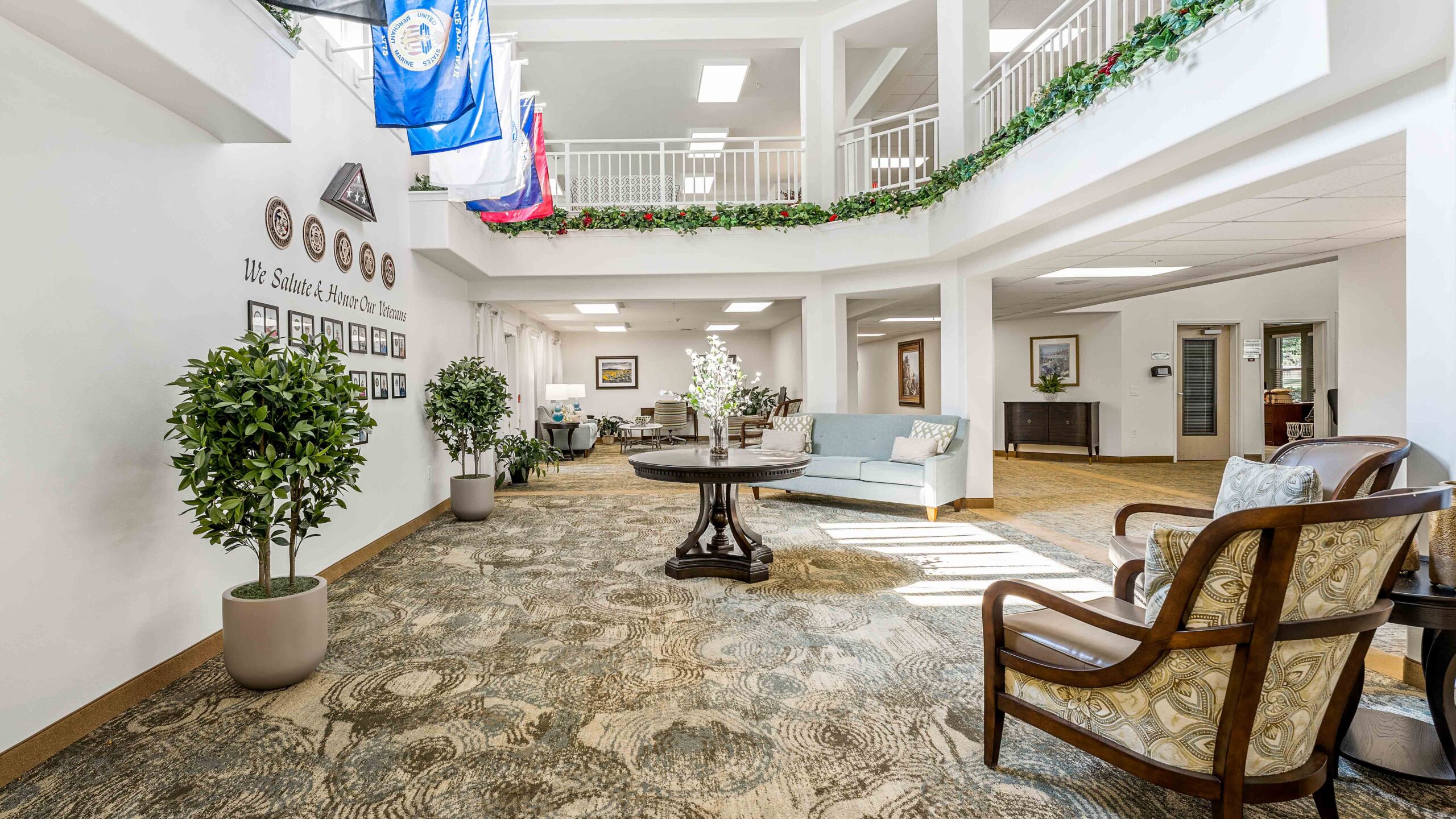Spacious, well-lit lobby with comfortable seating, indoor plants, and a round table with flowers. Flags and framed photos decorate the white walls. An upstairs railing is visible above.